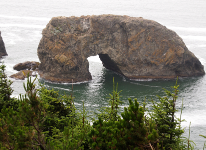 [A rectangular shaped rock near the edge of the evergreen-treed shore. This rock has a flat top and a curved notch at its bottom extending nearly halfway up the rock through which water flows.]